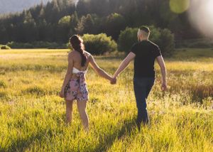 A couple holds hands while walking through a sunlit green meadow with mountains and trees in the background.