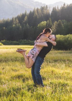 A man lifts and spins a woman in a floral dress in a sunny mountain meadow surrounded by tall trees.