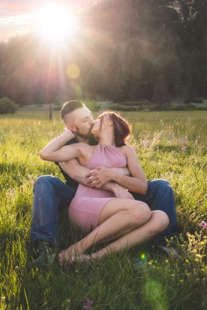 Couple sharing a romantic kiss while sitting in a sunlit meadow with golden hour light streaming behind them.