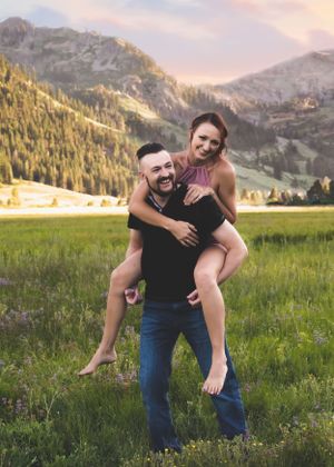 Man giving woman a piggyback ride while both smile in a beautiful mountain meadow setting.