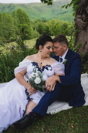 Bride and groom sitting together under a tree with lush green mountain backdrop on their wedding day.