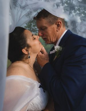 Groom tenderly lifting bride's chin for a kiss under a flowing white veil outdoors.