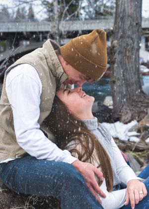 Couple sharing romantic kiss while sitting outdoors in falling snow during winter engagement session.