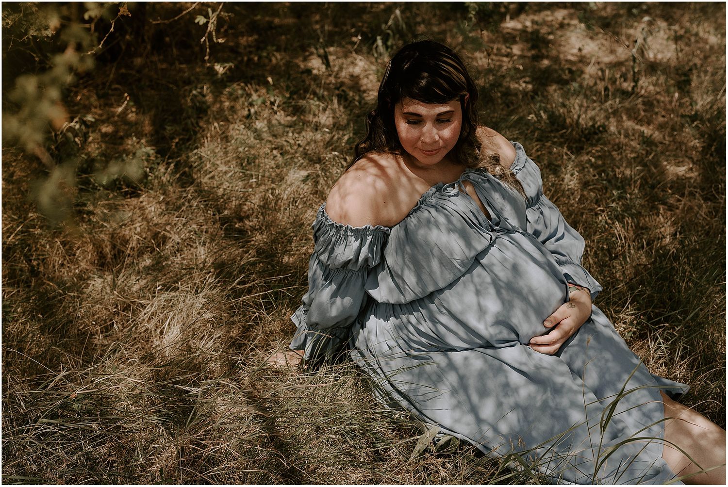 Dreamy maternity portrait with sunlight casting dappled shadows on a grey dress in nature.