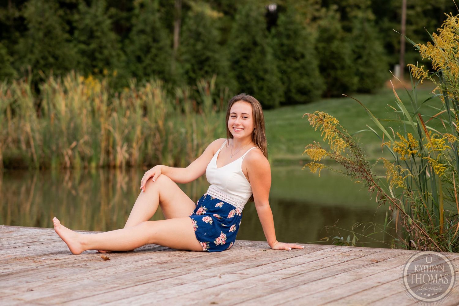 A person relaxes on a wooden dock by a pond with cattails and autumn foliage in the background.