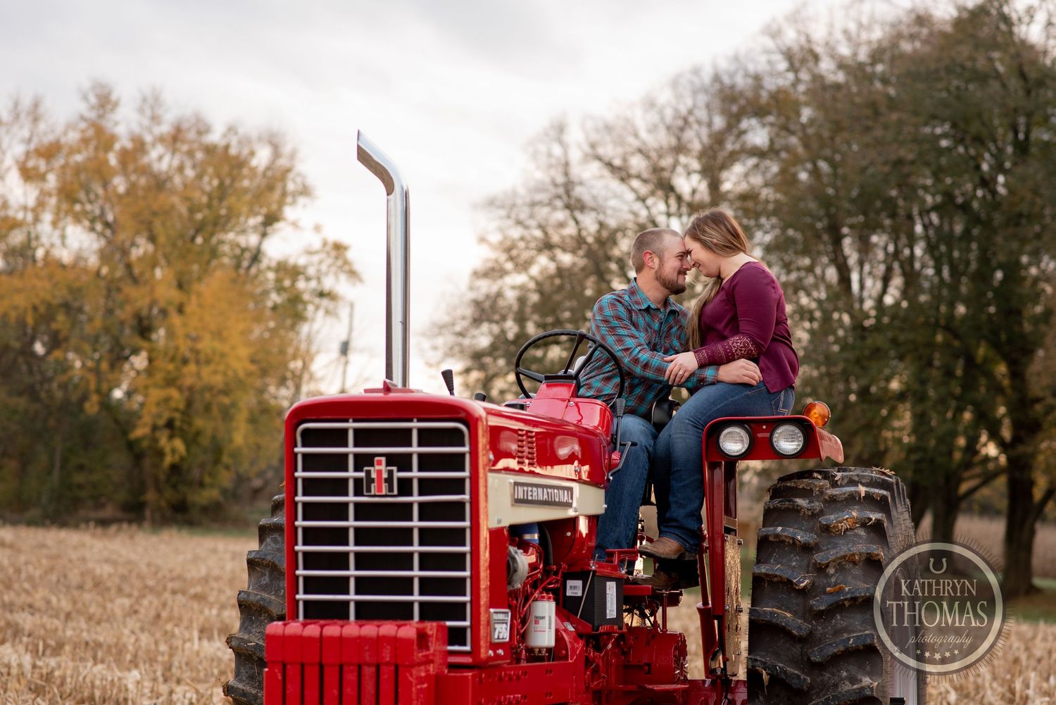 A couple sits together on a vintage red tractor surrounded by fall foliage.
