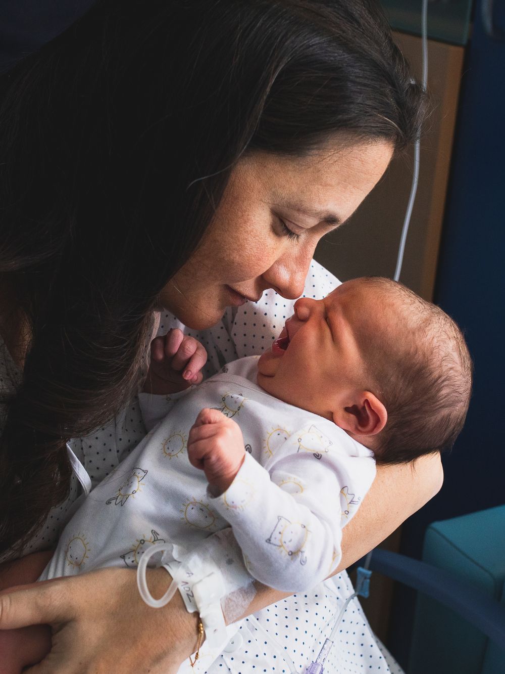 Mother in hospital gown holds and kisses newborn baby while partner looks on.