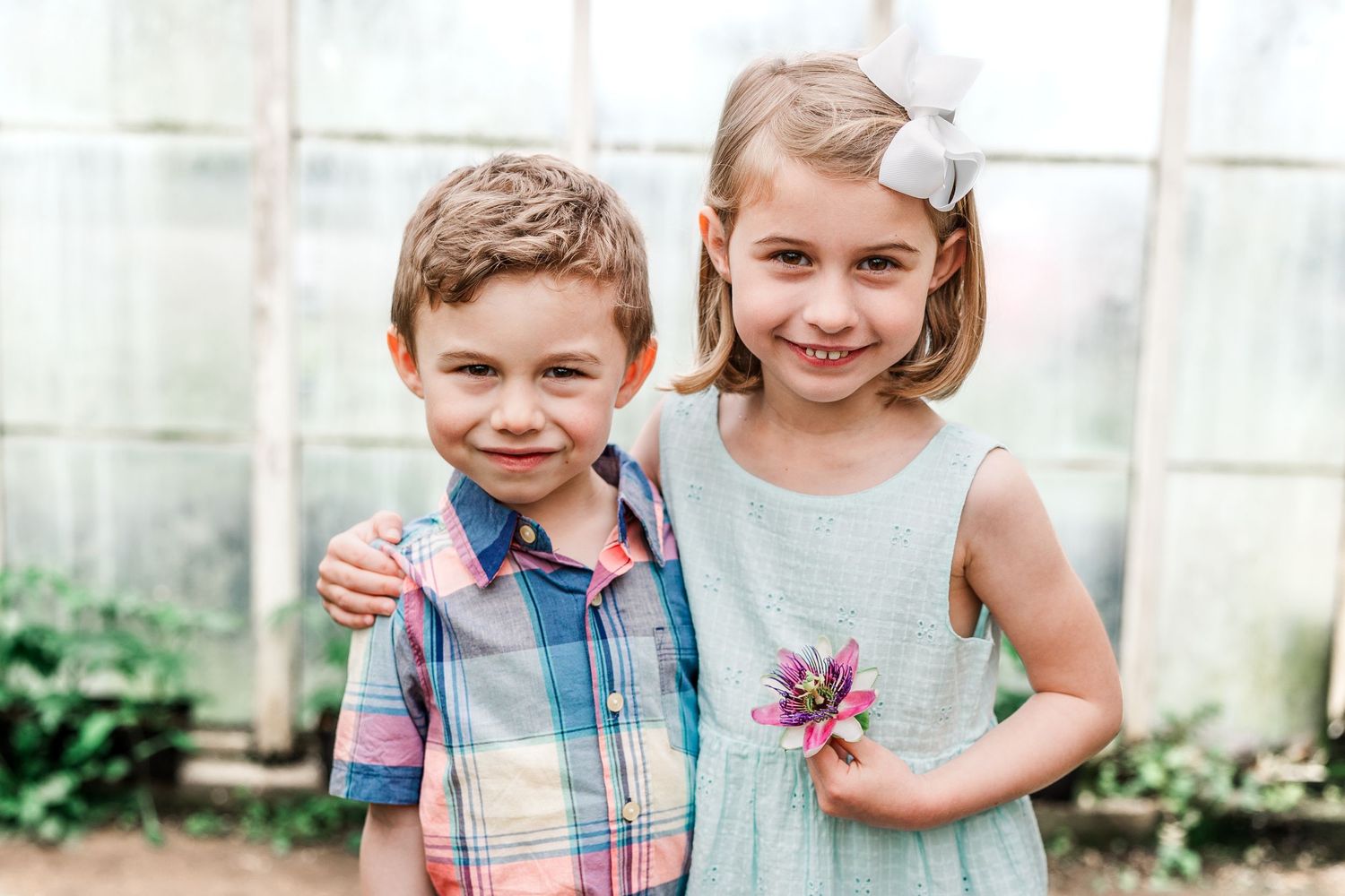 Two young figures pose together in a greenhouse with plants in the background.