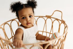 Small child sitting in rattan chair wearing white dress against neutral background.