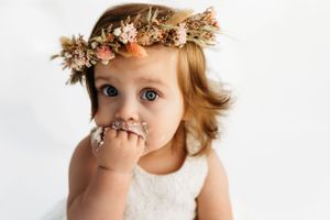 A delicate floral crown adorns light strawberry blonde hair contrasting with a white dress against a soft white backdrop.