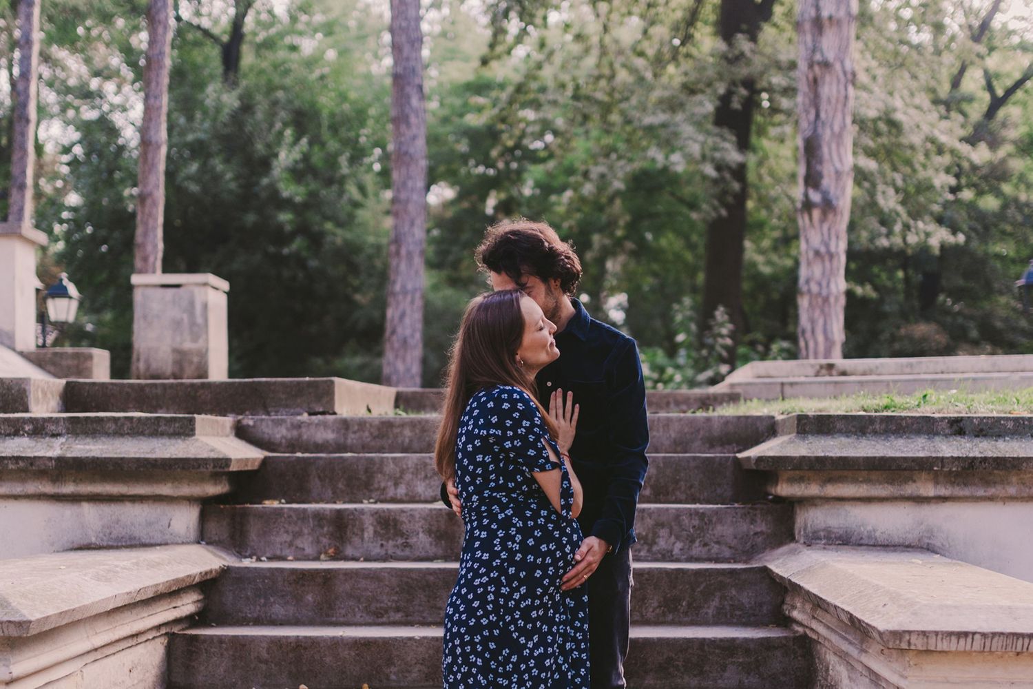 A romantic couple embraces on stone steps surrounded by tall trees in a park setting.