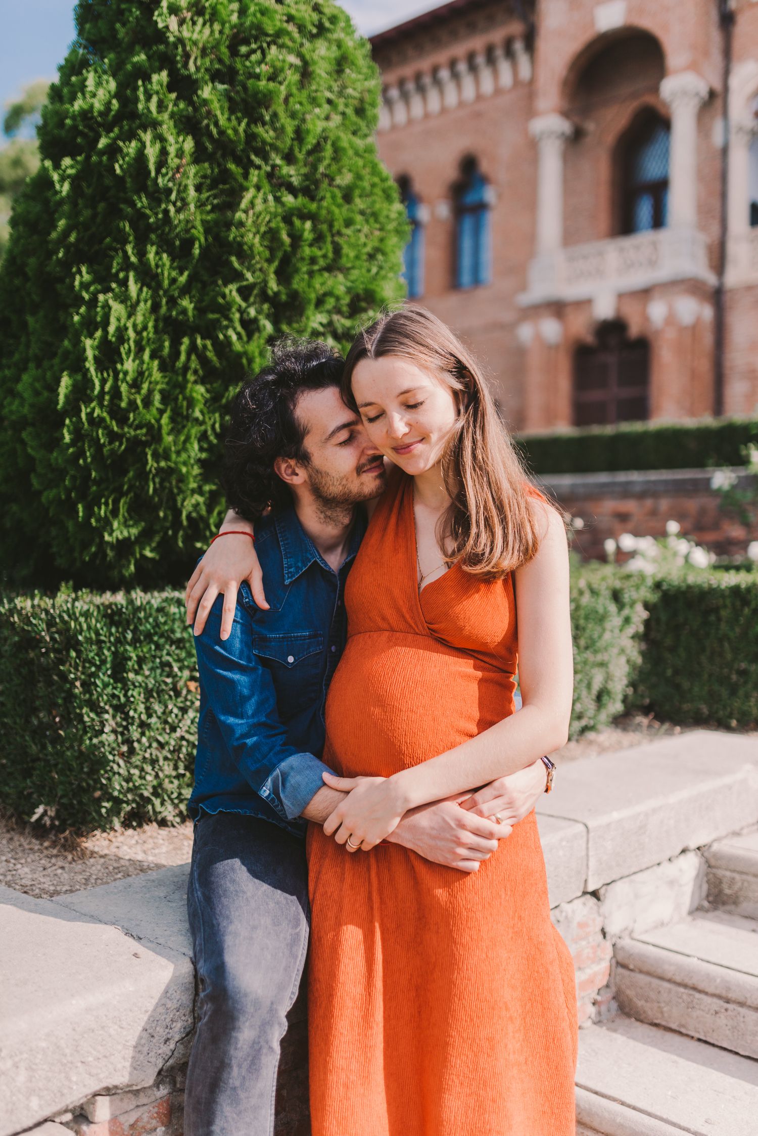 Expecting couple poses for photos in garden setting with historic brick building backdrop.