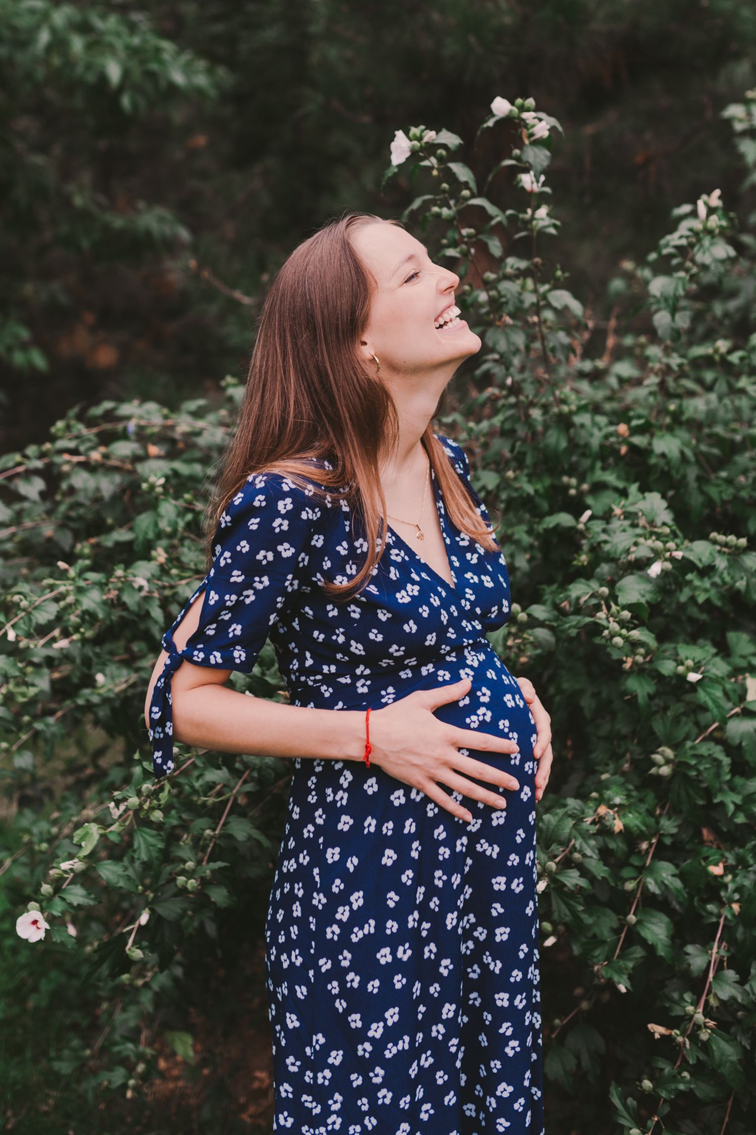 A pregnant woman in a navy floral dress laughs joyfully while standing in front of green foliage.