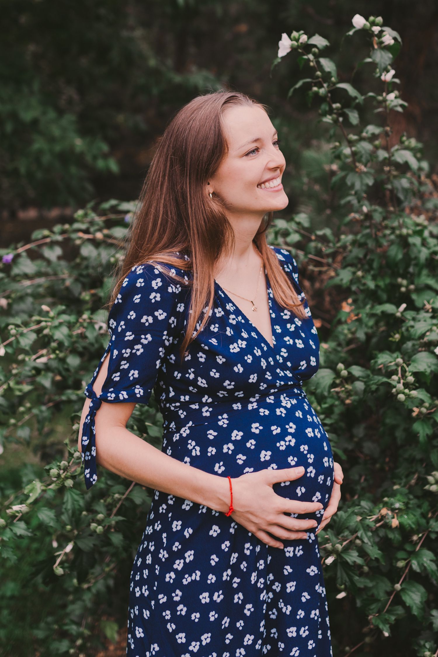 A woman in a blue floral dress embraces her baby bump while laughing outdoors.