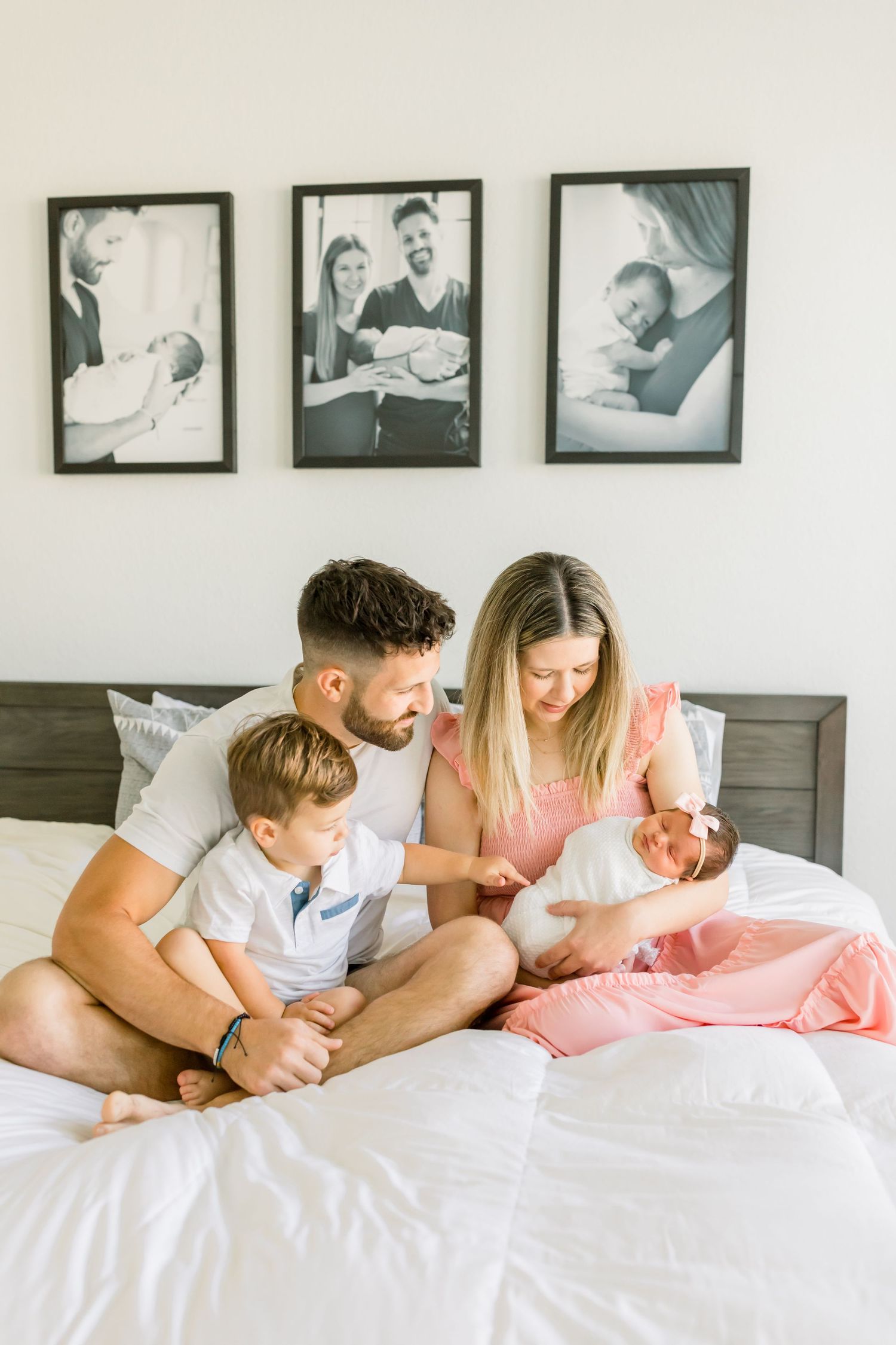 A family sits together on a white bed with framed photos on the wall above them.