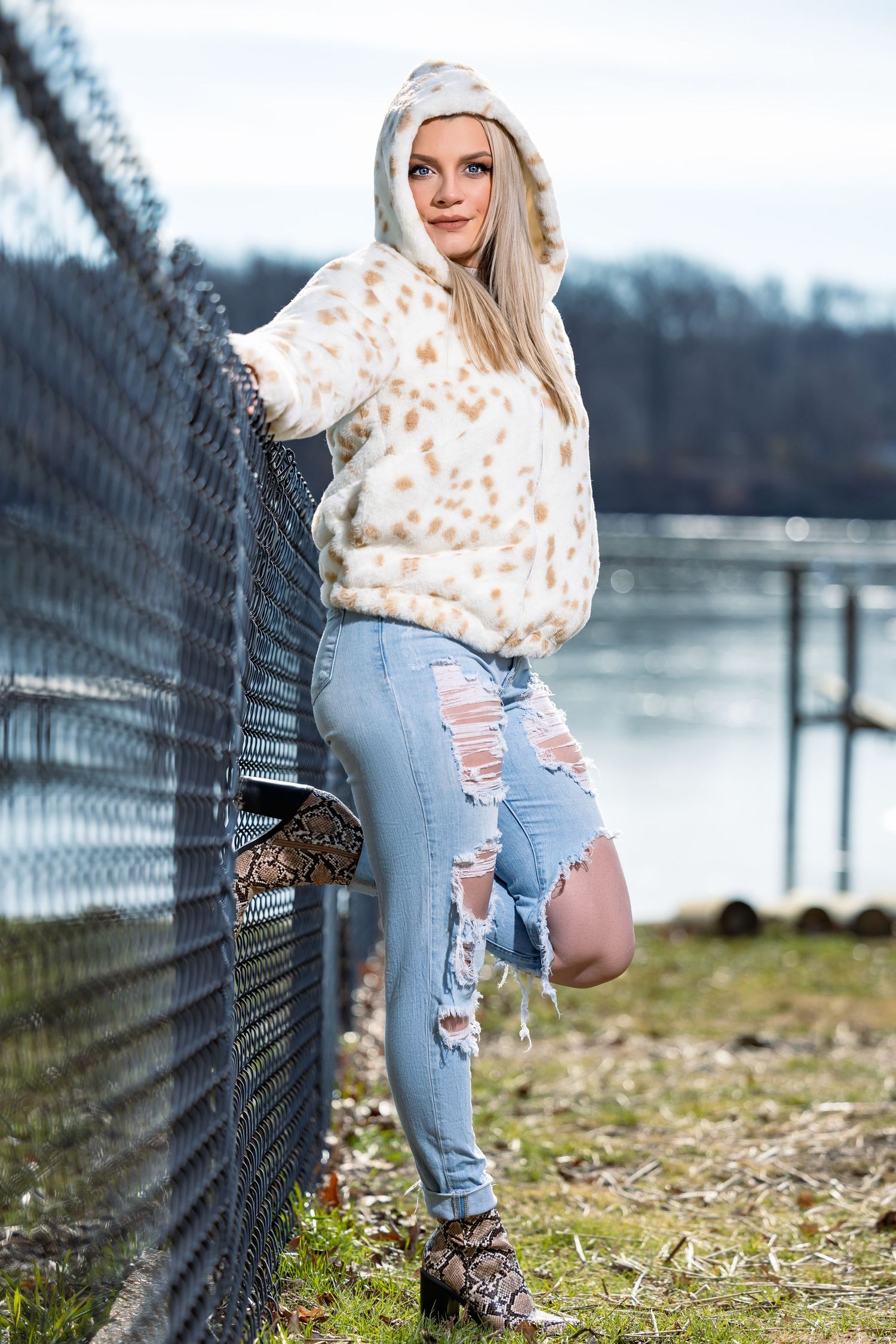 Woman in leopard print hoodie and distressed jeans leans against fence by river.