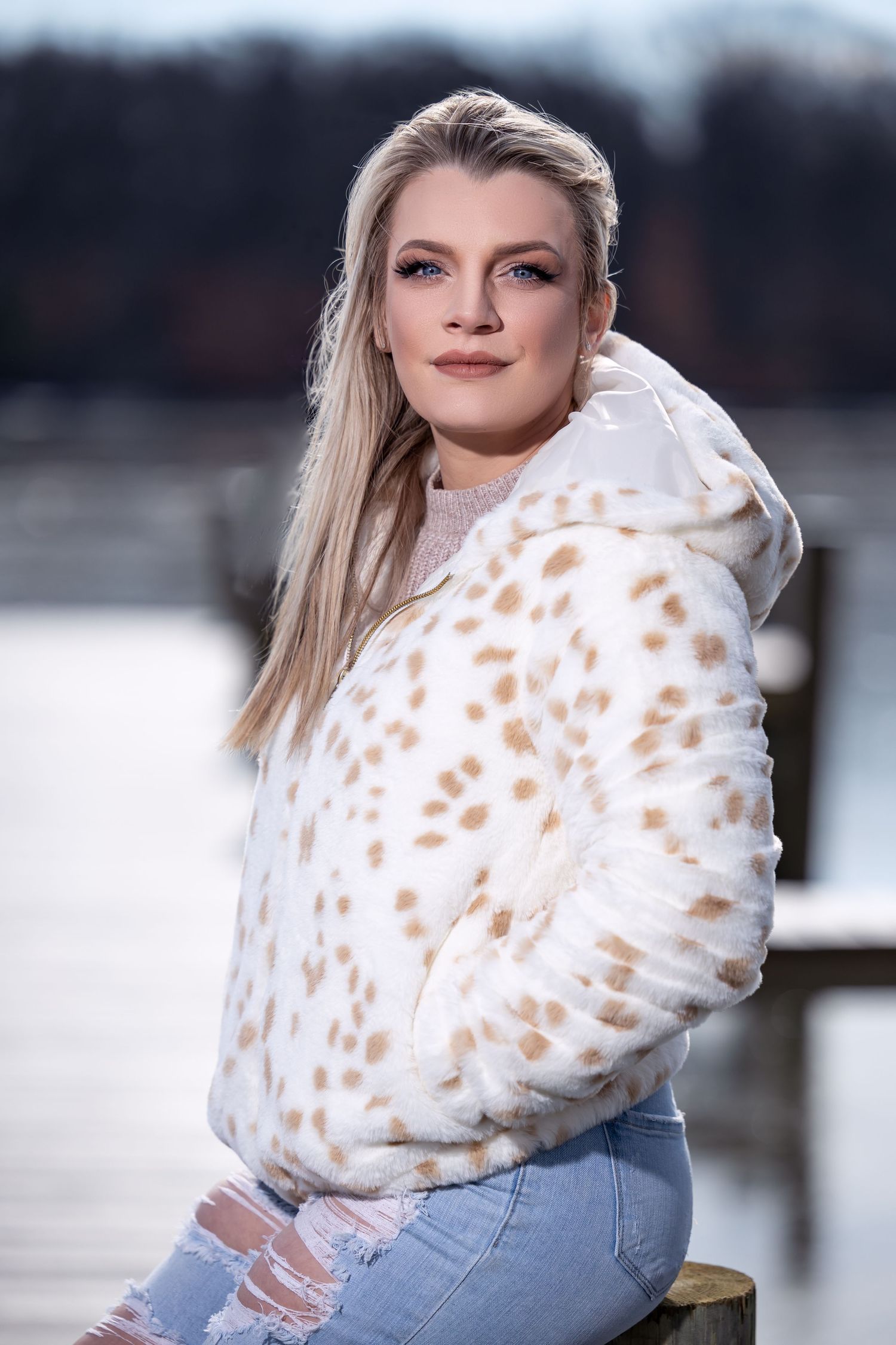 Woman in cream spotted jacket and ripped jeans poses outdoors by water.