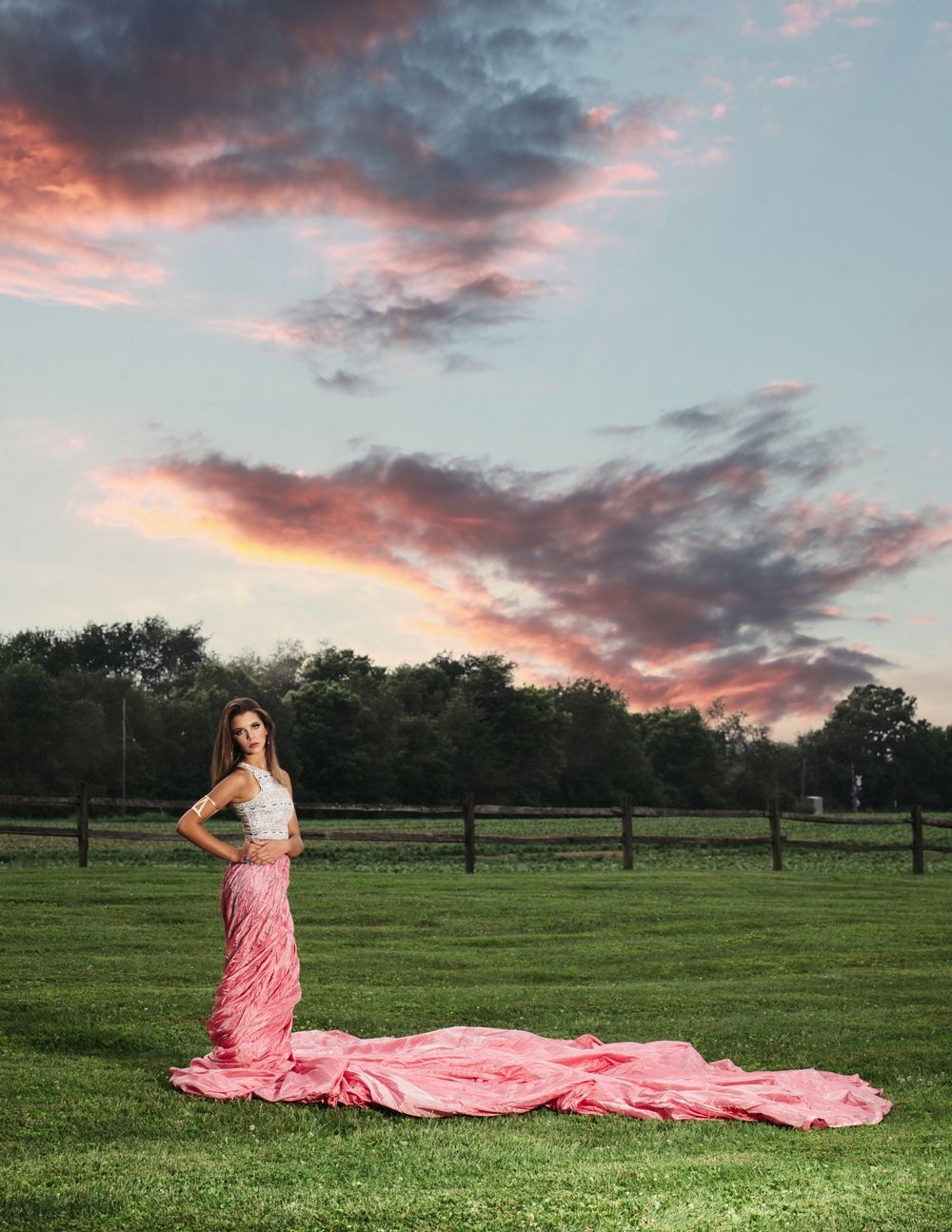 Dramatic sunset portrait in field with flowing pink dress against colorful sky.