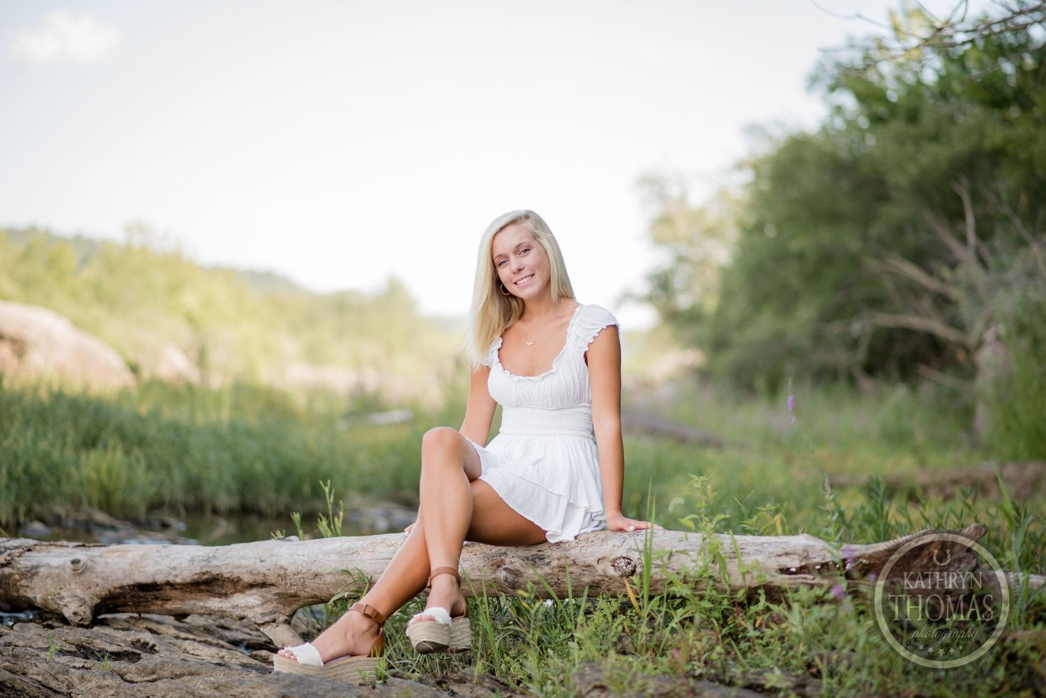 Woman in white dress sitting on rustic log by peaceful waterside.