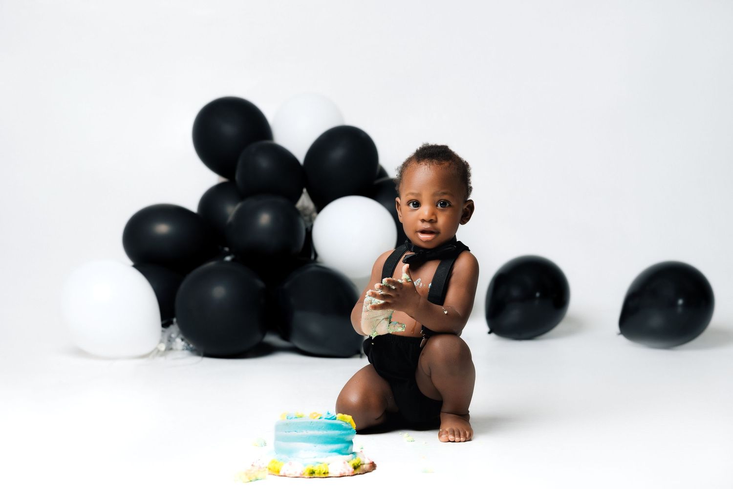 Adorable baby boy celebrating first birthday with smash cake surrounded by black and white balloons.