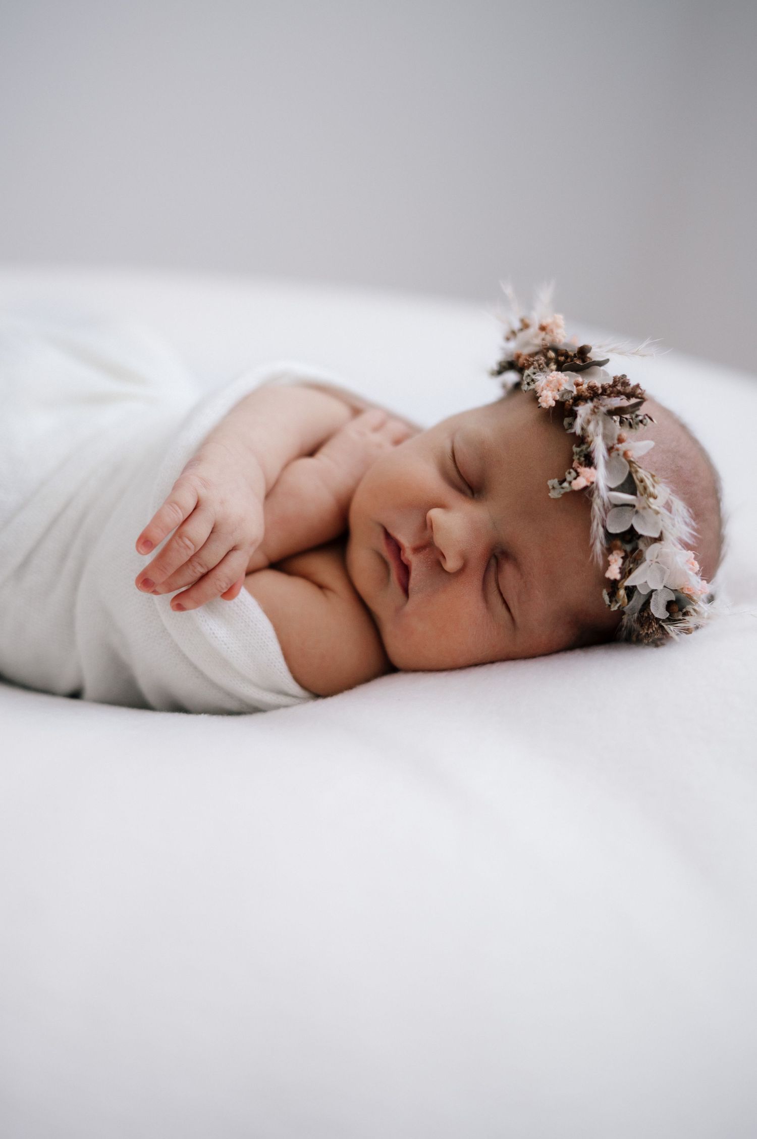 Newborn wearing floral crown and white wrap sleeping peacefully on white surface.