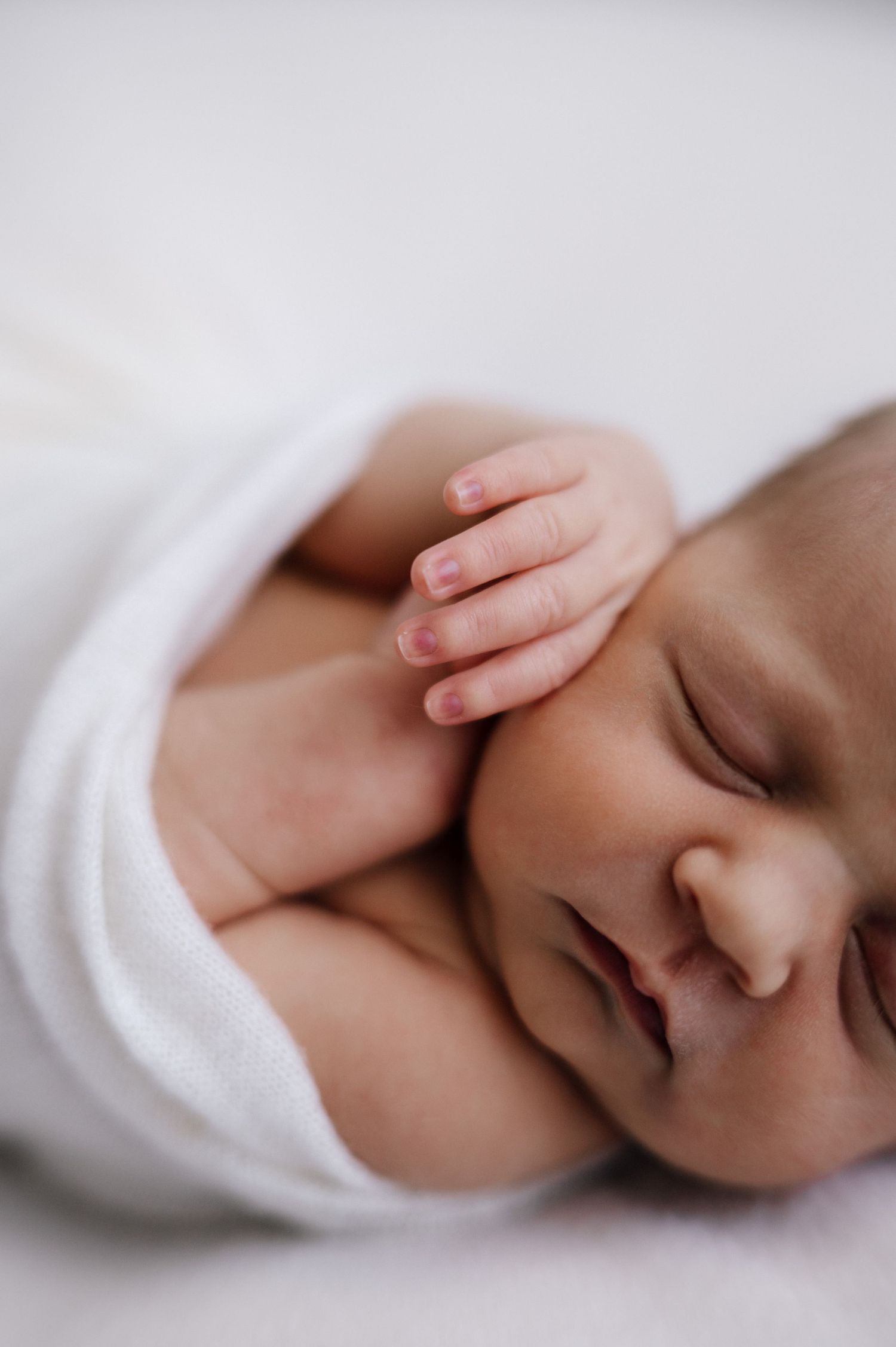 Close-up of sleeping baby wrapped in white cloth with tiny hands visible.