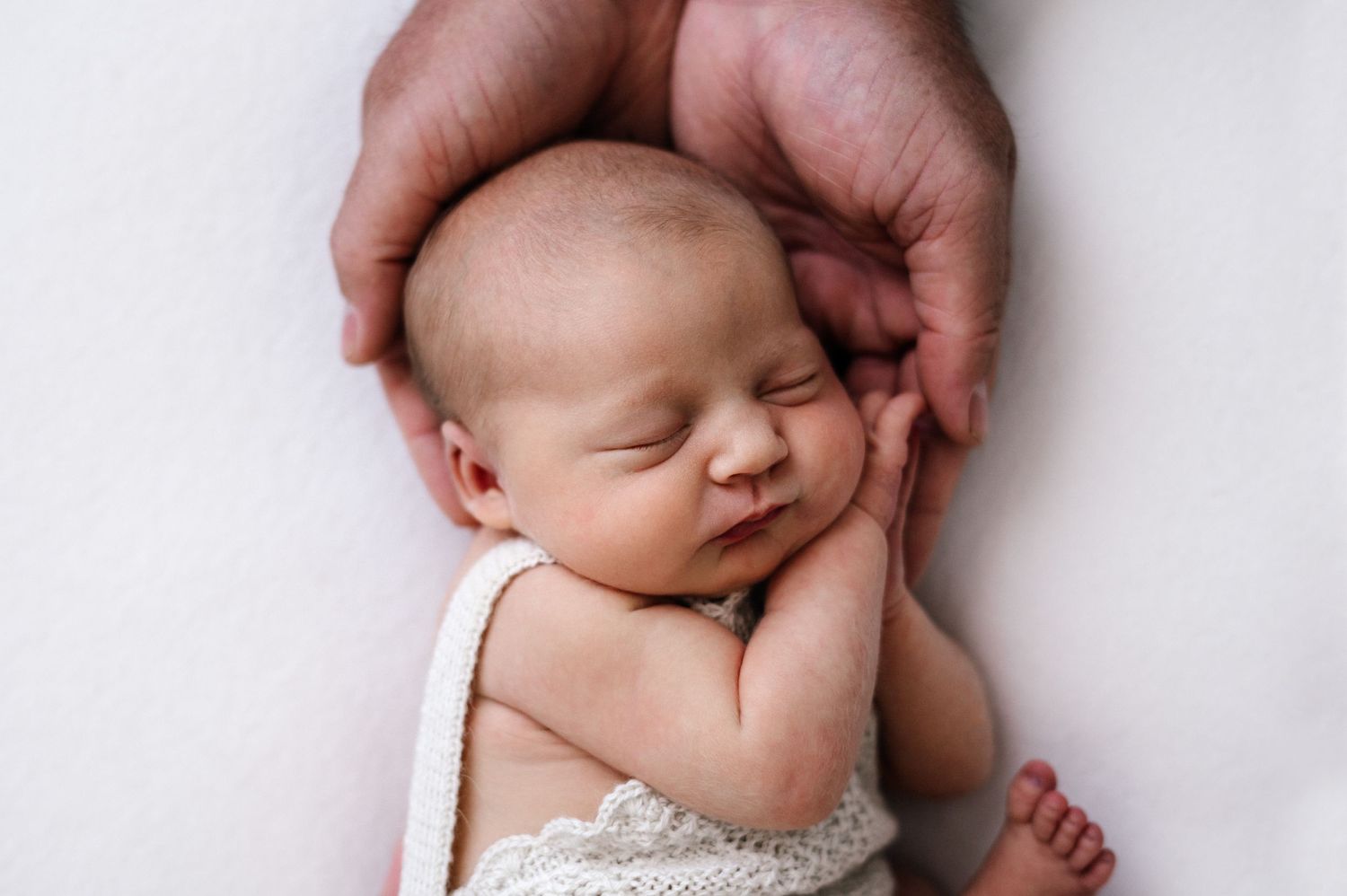 Sleeping newborn in lace outfit cradled by caring hands on white background.