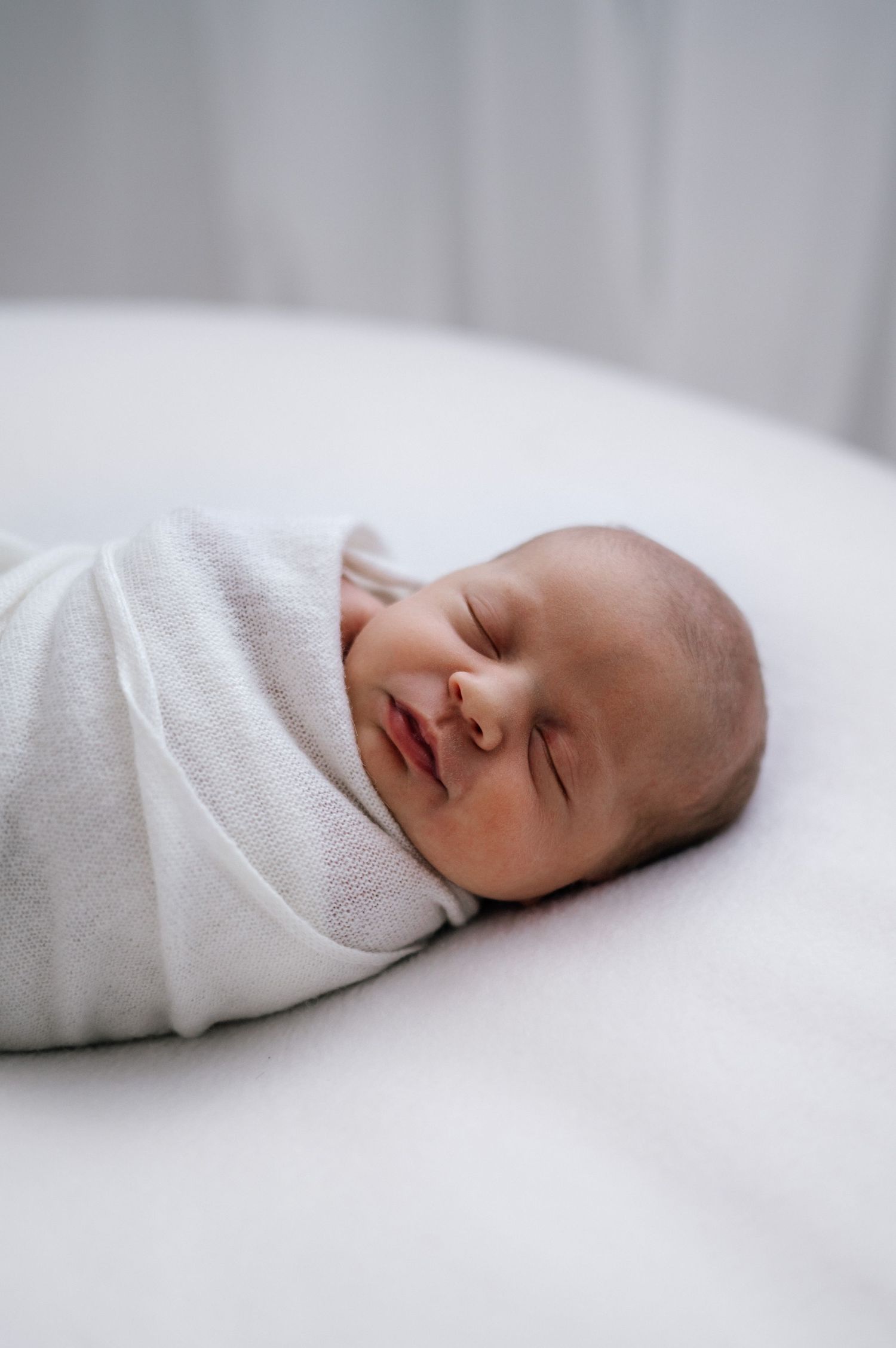 Sleeping newborn wrapped snugly in white swaddle on light colored surface.