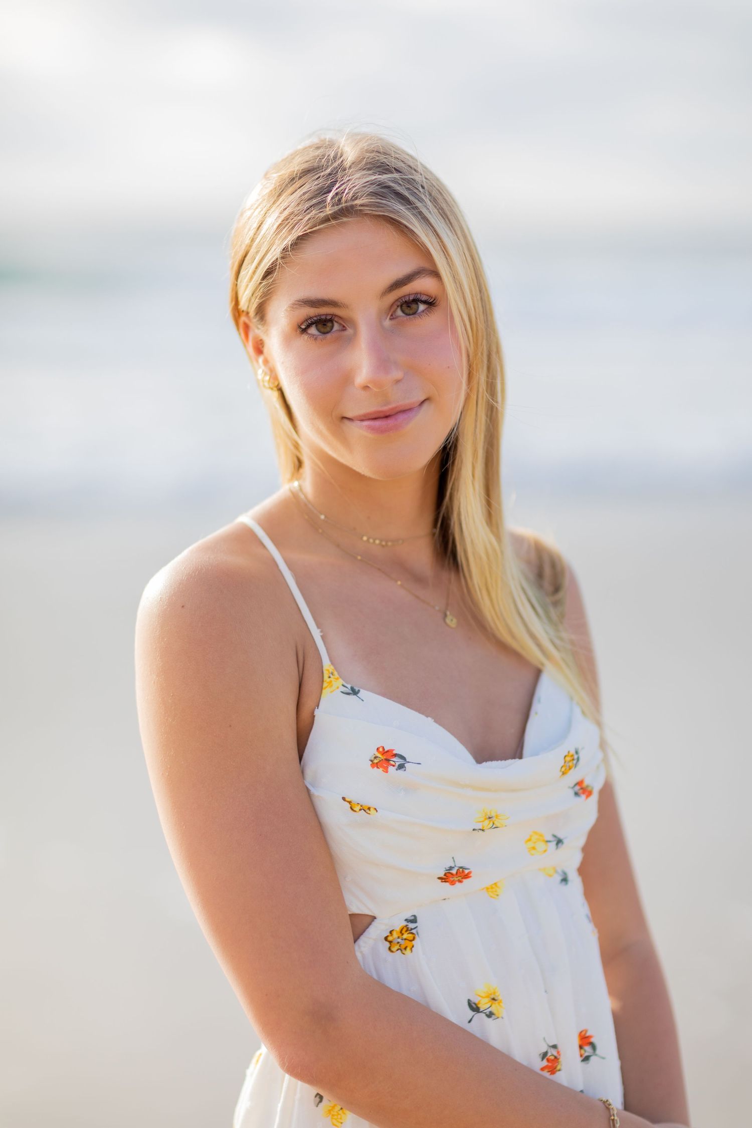 A high school senior in white floral sundress poses at beach during golden hour photography session.