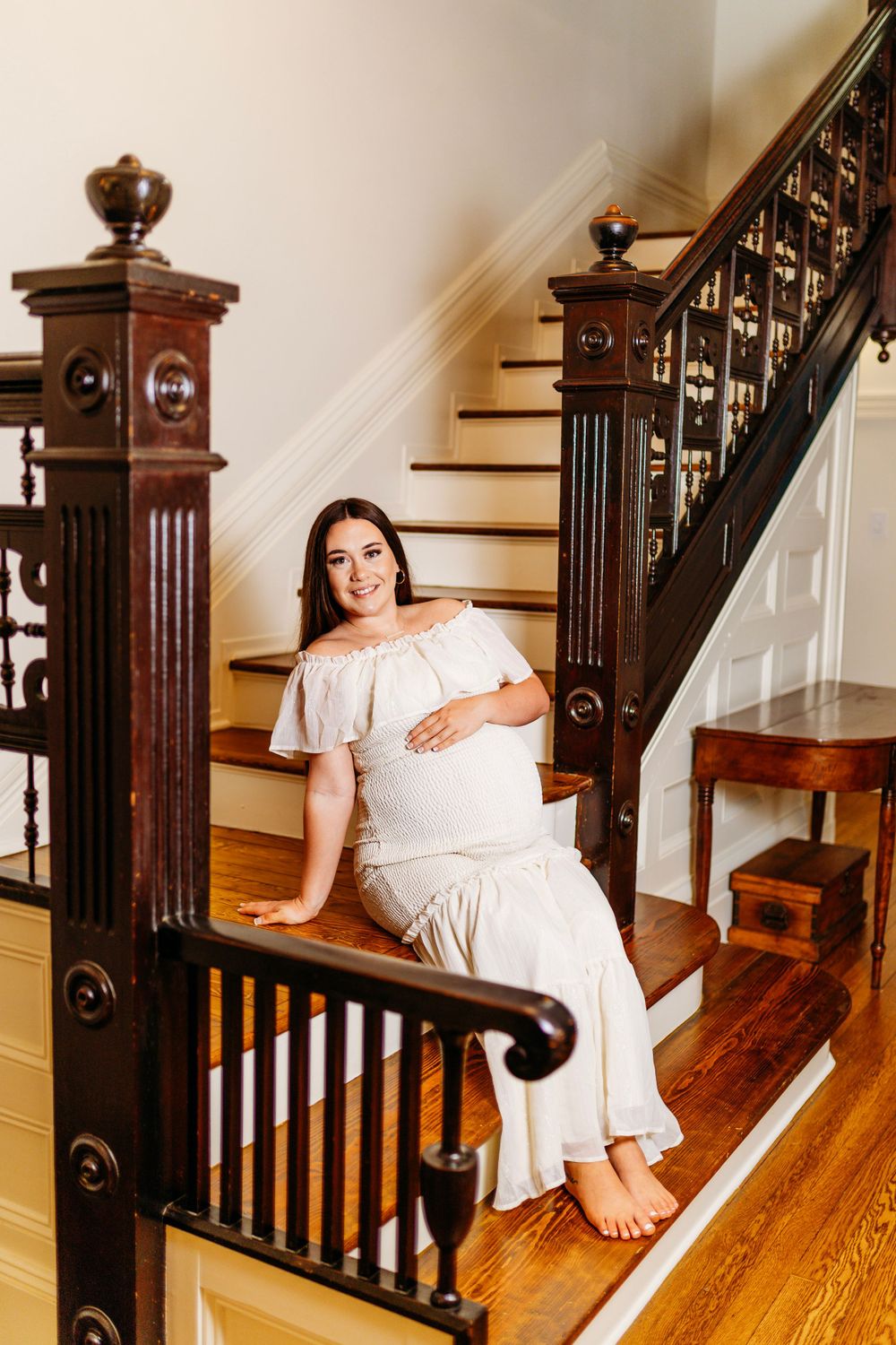 Smiling pregnant woman in white off-shoulder dress sitting barefoot on elegant wooden staircase.