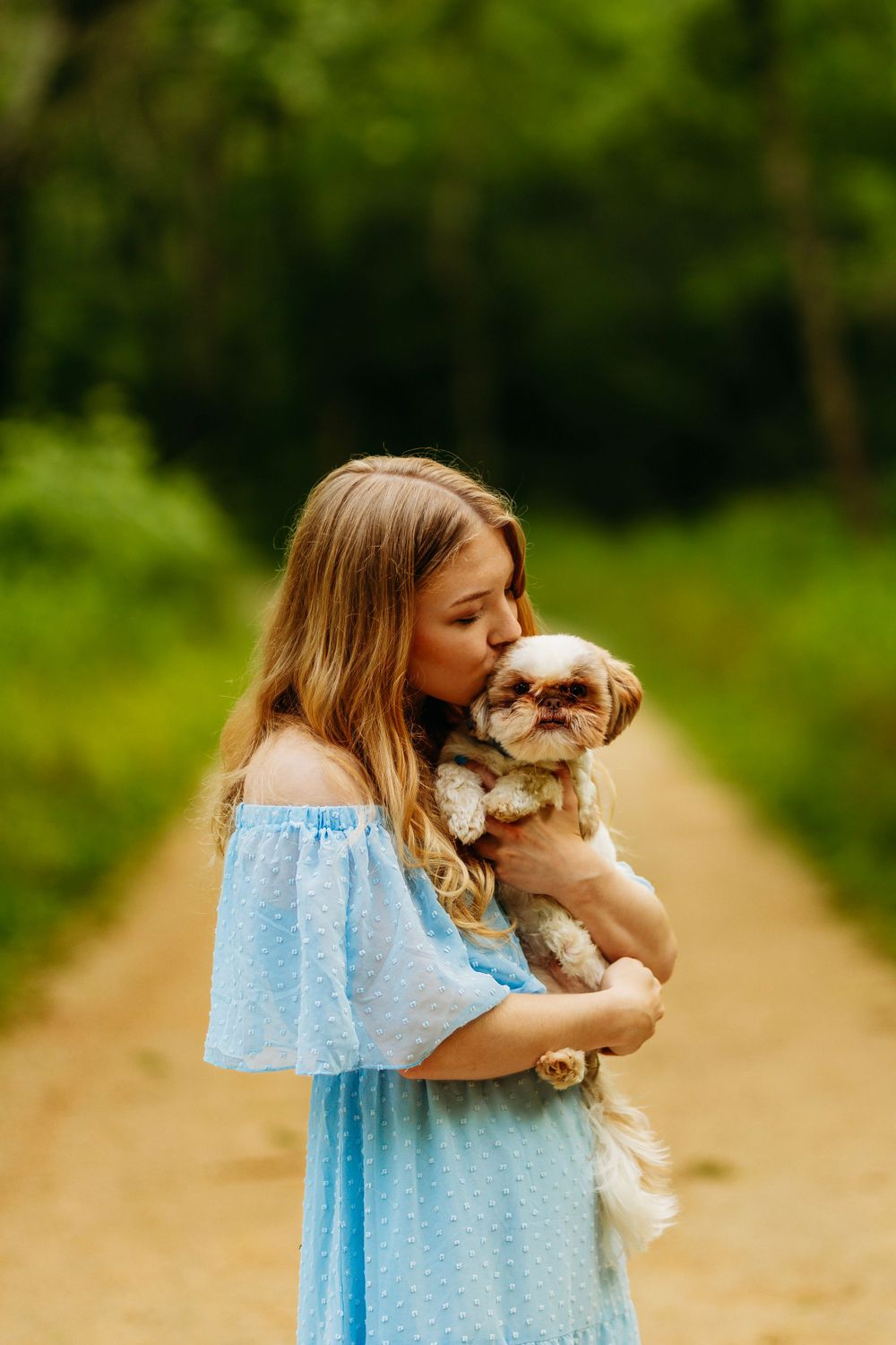 A young blonde woman in a blue off-shoulder dress kissing her Shih Tzu dog on a forest path.