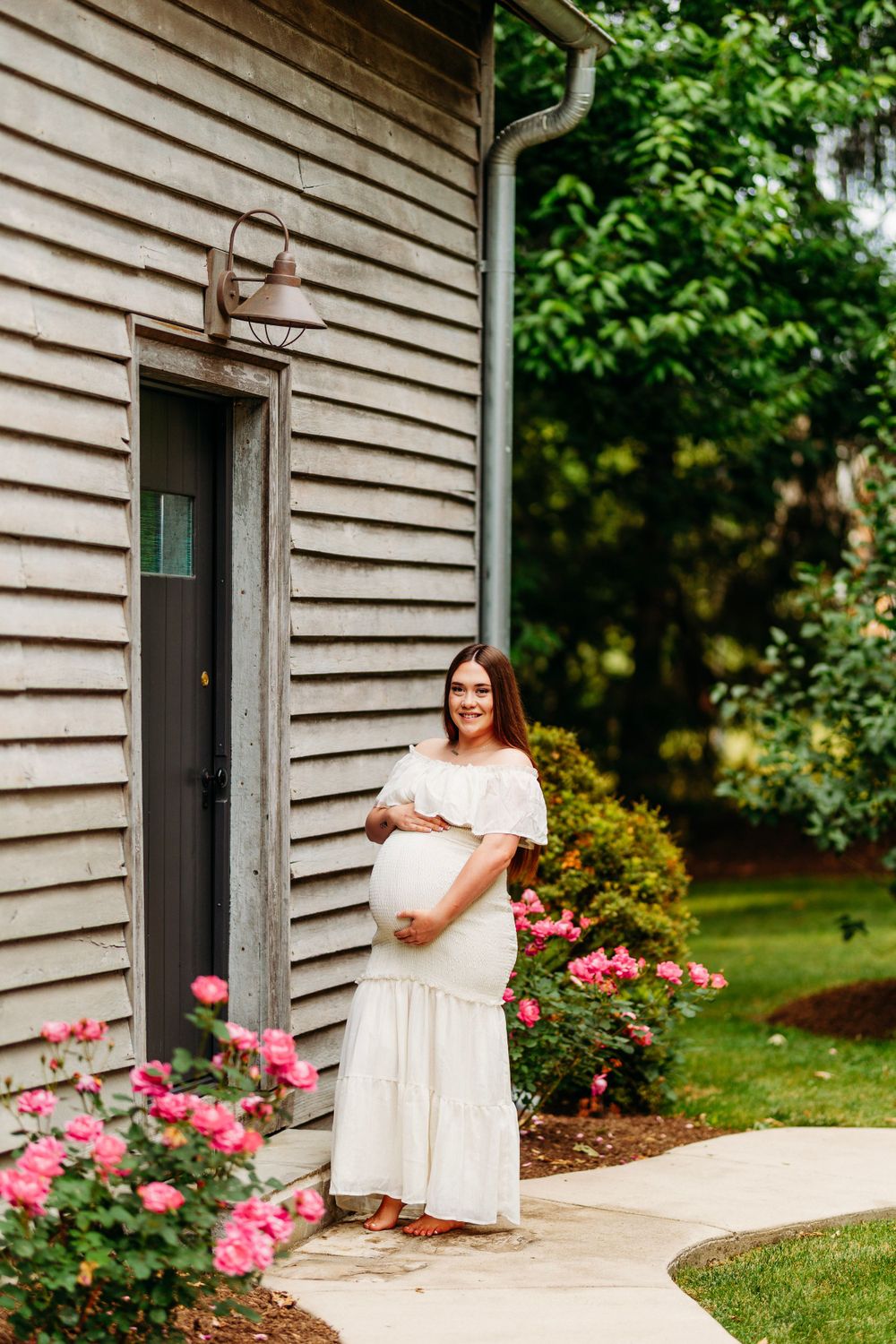 Smiling pregnant woman in white off-shoulder dress holding baby bump beside pink roses near rustic wooden building.