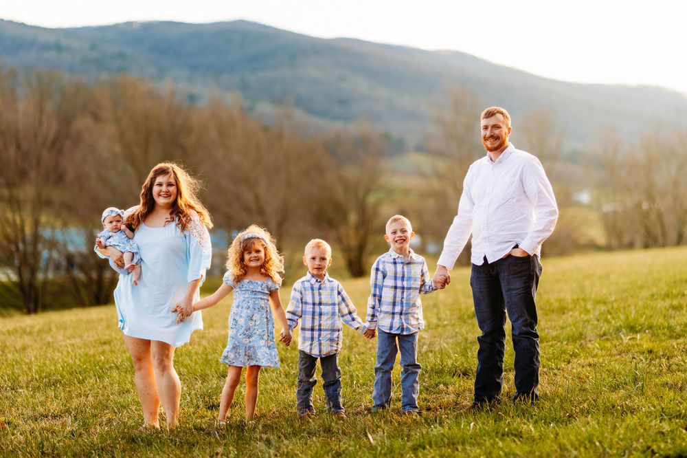 Happy family of six poses holding hands in a green field with mountain backdrop at golden hour.
