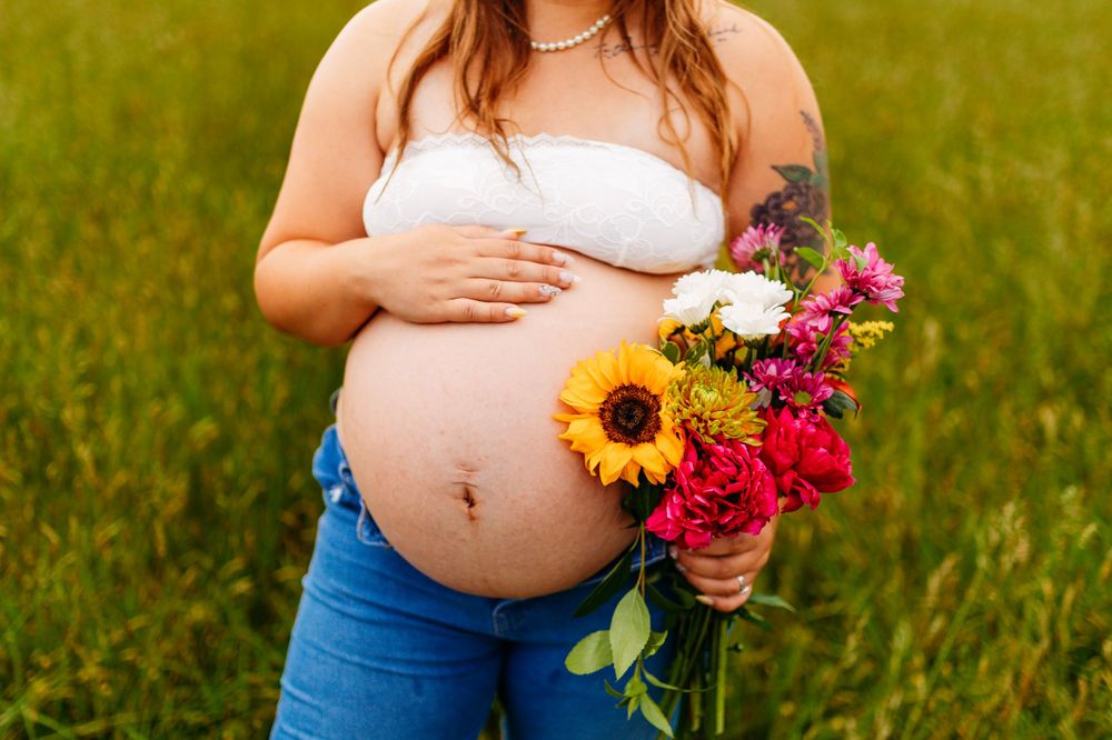Close-up of pregnant belly with colorful sunflower bouquet in a green field setting.