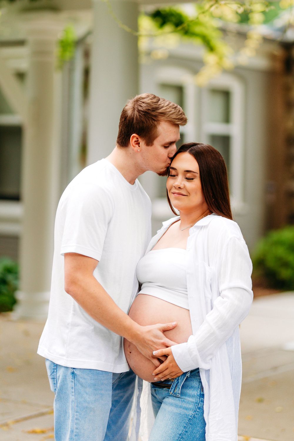 Man kissing pregnant woman's forehead while both cradle her bare baby bump outdoors in soft natural light.