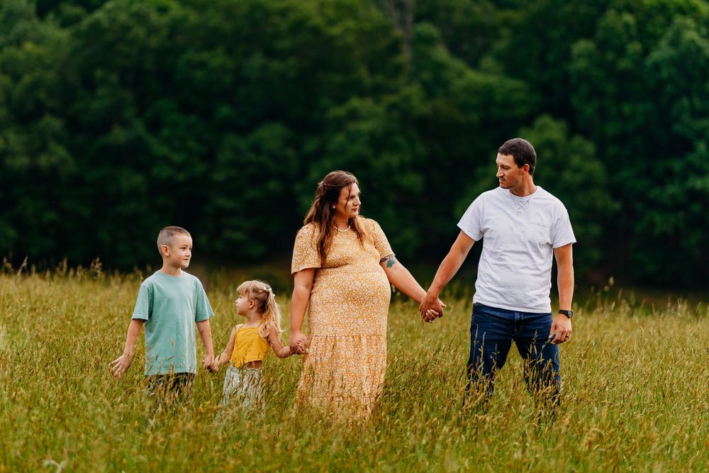 Happy pregnant family of four holding hands walking through tall green grass field with forest backdrop.