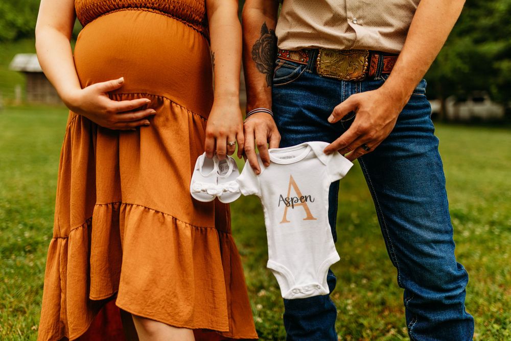 Couple holding personalized baby onesie with letter A outdoors during rustic country maternity photo session.