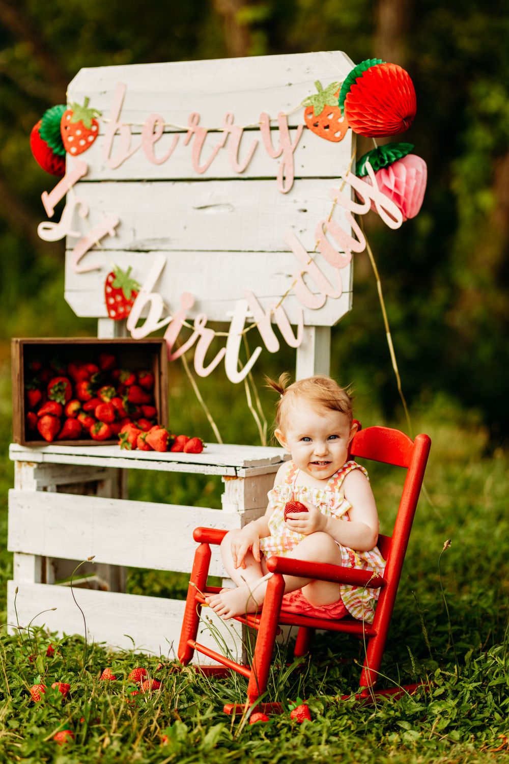 Smiling baby in floral outfit sits on red rocking chair at strawberry-themed first birthday setup.