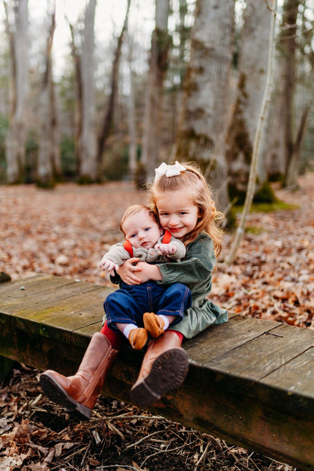 Young girl hugging baby sibling while sitting on a wooden bench in autumn woods.