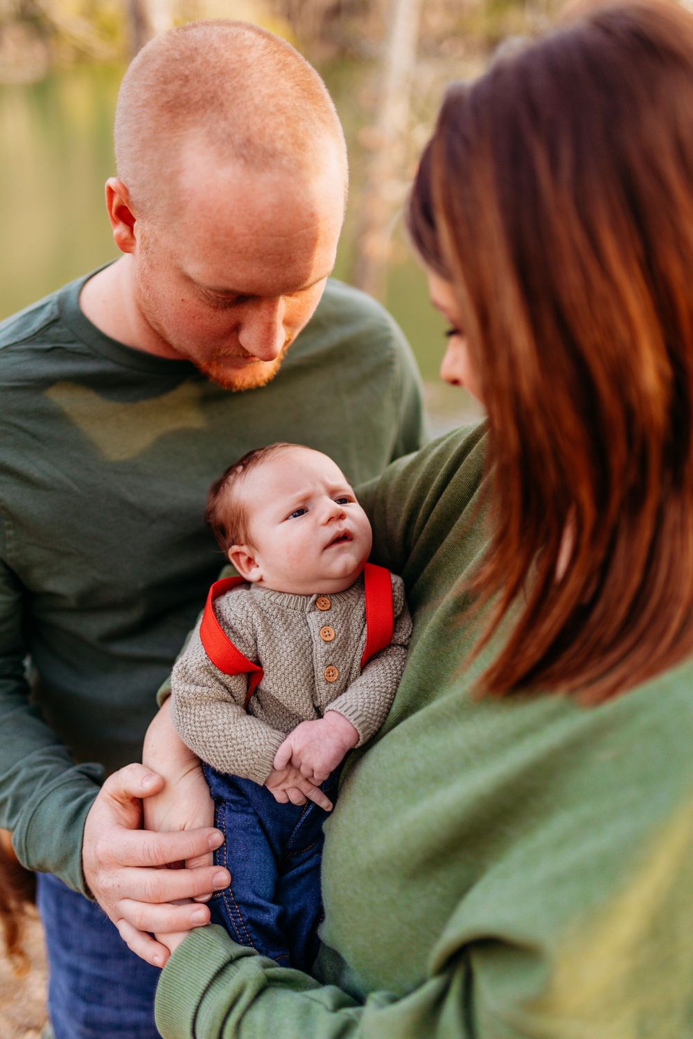 New parents in green sweaters lovingly looking down at their alert newborn baby outdoors in fall.