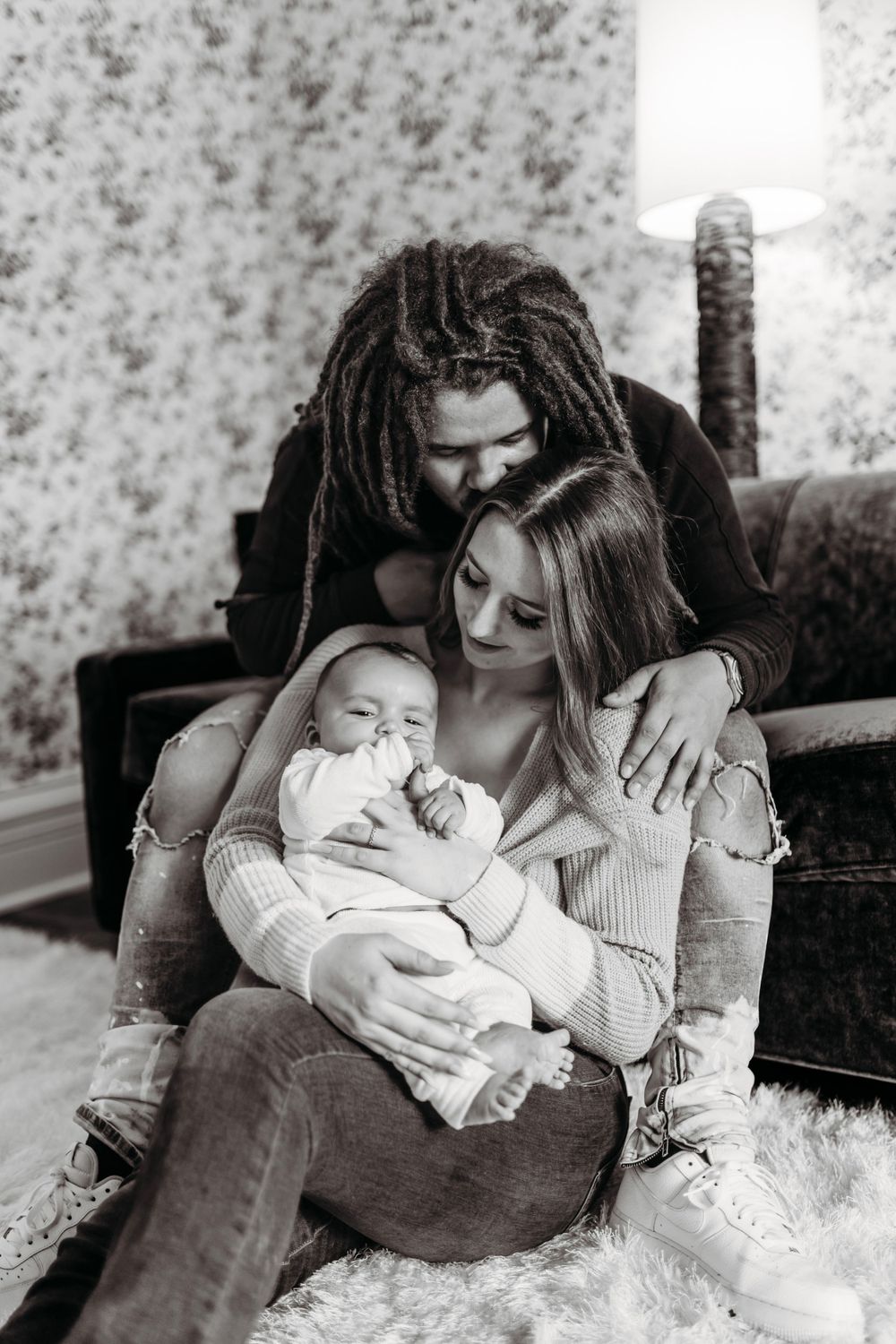 Black and white portrait of interracial couple tenderly holding sleeping newborn baby on white fur rug.