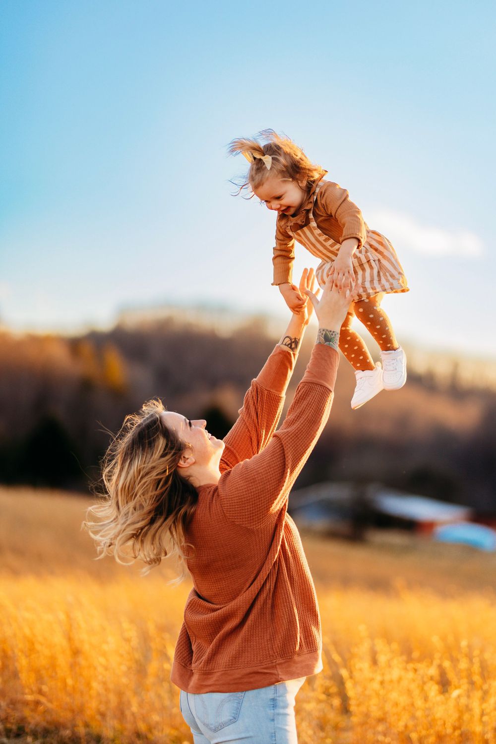 Joyful mother lifting her laughing toddler daughter high in the air in a sunlit golden autumn field.