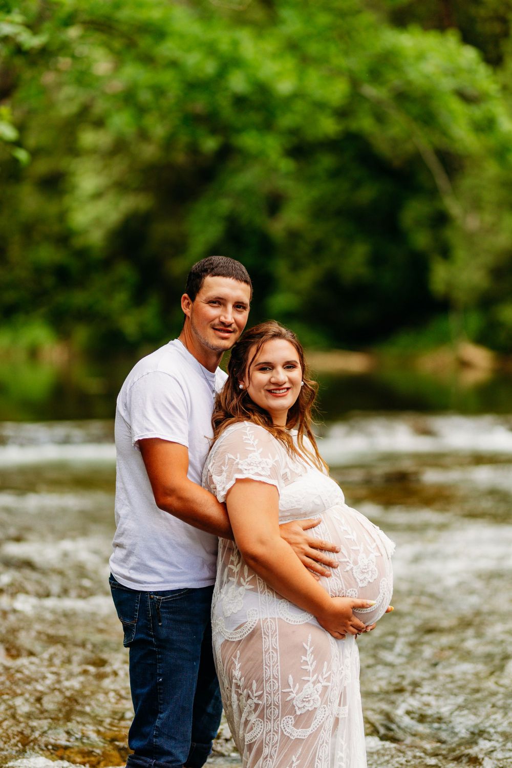 Couple posing for maternity photos by a rocky creek surrounded by lush green trees.