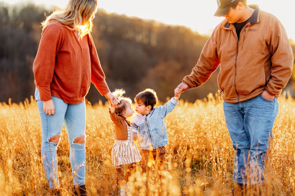 Family of four holding hands with two toddlers in a golden autumn field during warm sunset hour.