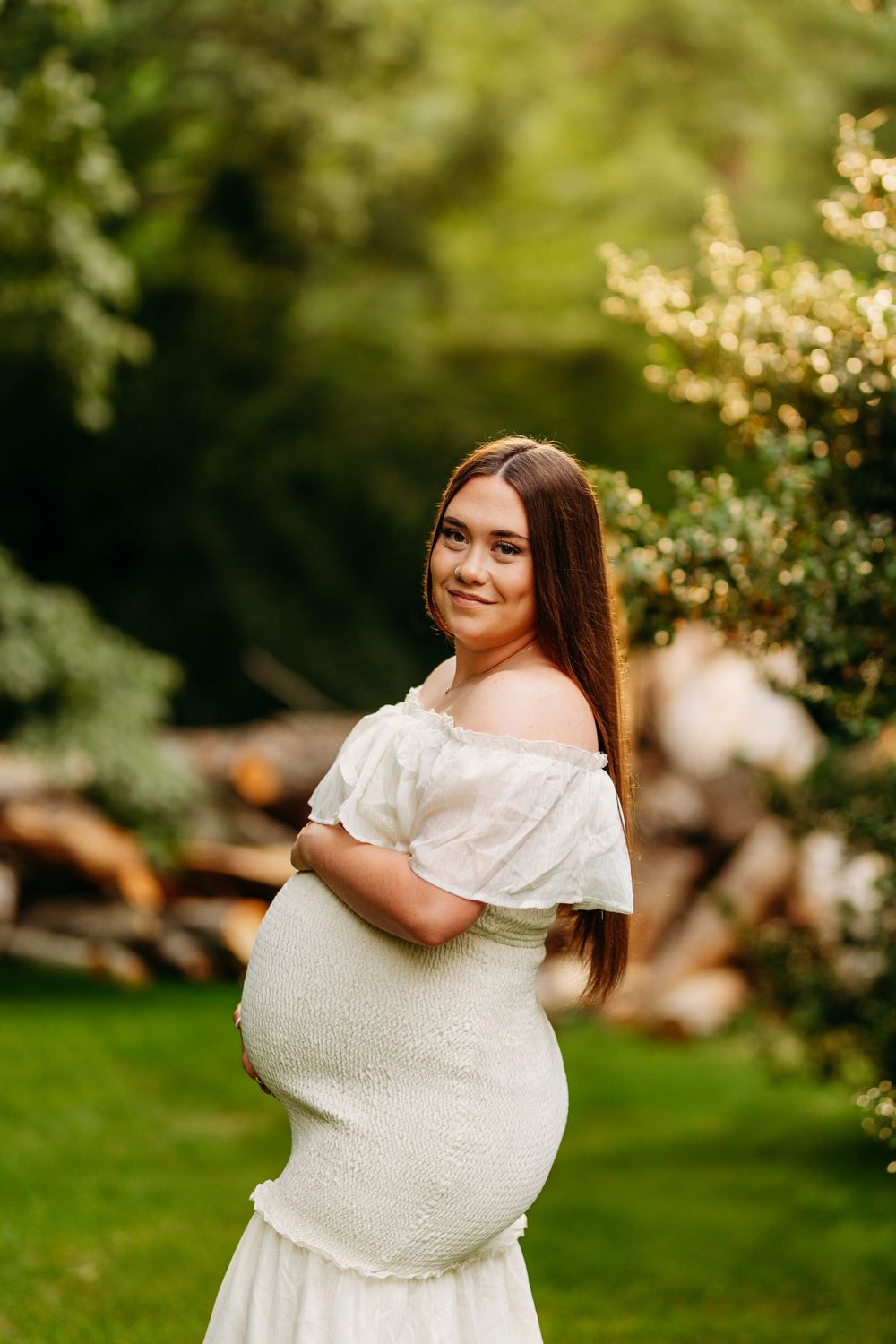 Smiling pregnant woman in white off-shoulder maternity dress poses outdoors in lush green garden setting.