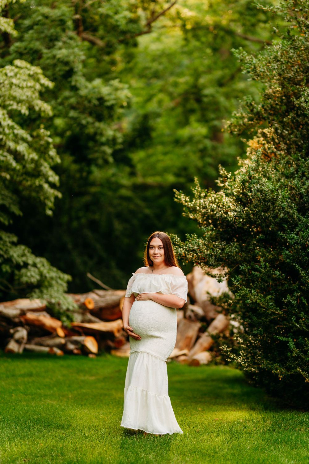 Pregnant woman in elegant white maternity gown stands gracefully among lush green trees outdoors.