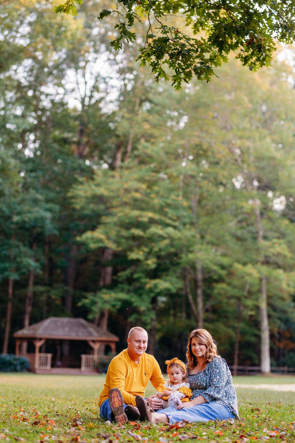 Happy family with toddler sits together on autumn grass in a lush green park with gazebo.