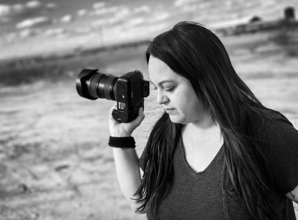 Photographer with long dark hair using professional camera against cloudy sky.