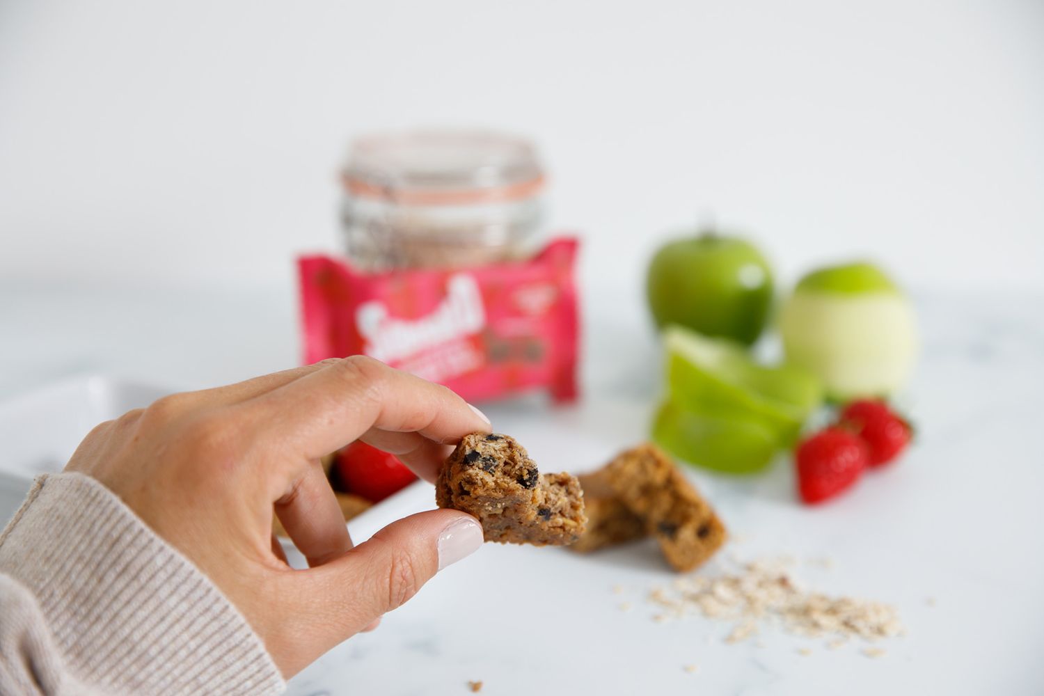 Hand reaching for a healthy granola bar with fresh fruit and nuts in the background from a product photoshoot.