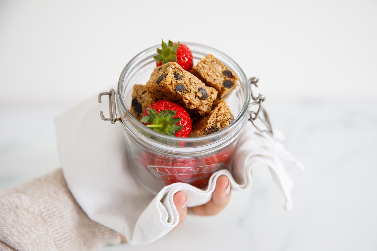 Glass jar filled with protein bars and fresh strawberries on white linen from a product photoshoot.